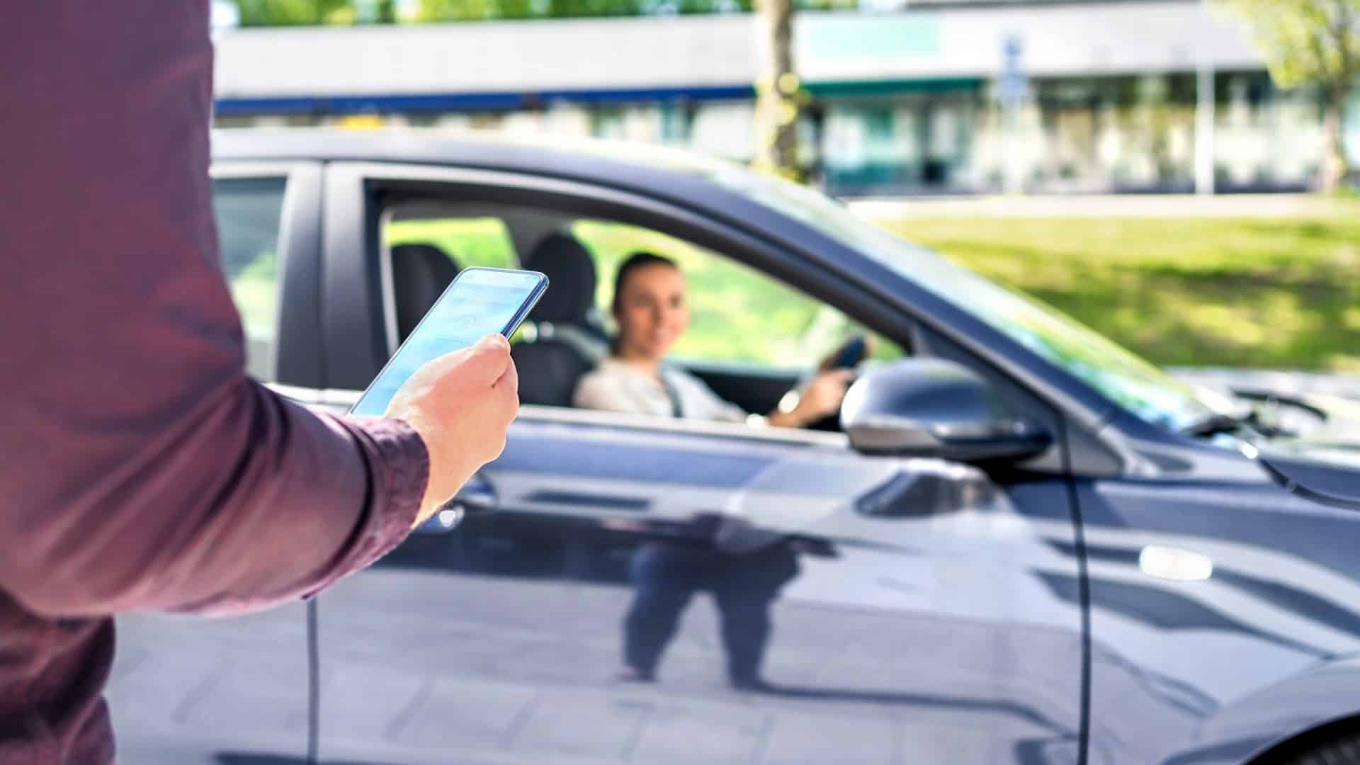 Em primeiro plano, uma pessoa com um smartphone na mão; no segundo plano, desfocado, uma mulher, sorrindo, dirigindo um carro preto