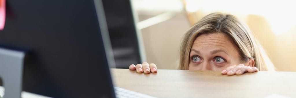 Mulher com os olhos assustados observando, da borda de uma mesa, um monitor de computador