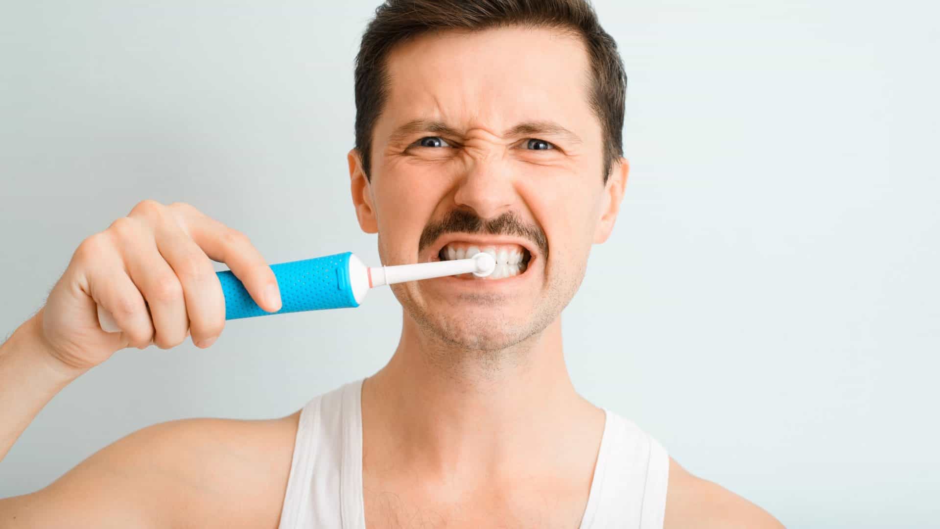 Retrato de um homem bonito escovando os dentes de forma persistente e enérgica com uma escova de dentes elétrica moderna, olhando para a câmera dentro de casa. A rotina diária de higiene bucal e clareamento dental de um jovem.