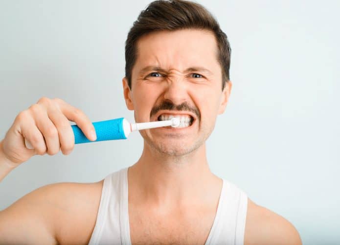 Retrato de um homem bonito escovando os dentes de forma persistente e enérgica com uma escova de dentes elétrica moderna, olhando para a câmera dentro de casa. A rotina diária de higiene bucal e clareamento dental de um jovem.