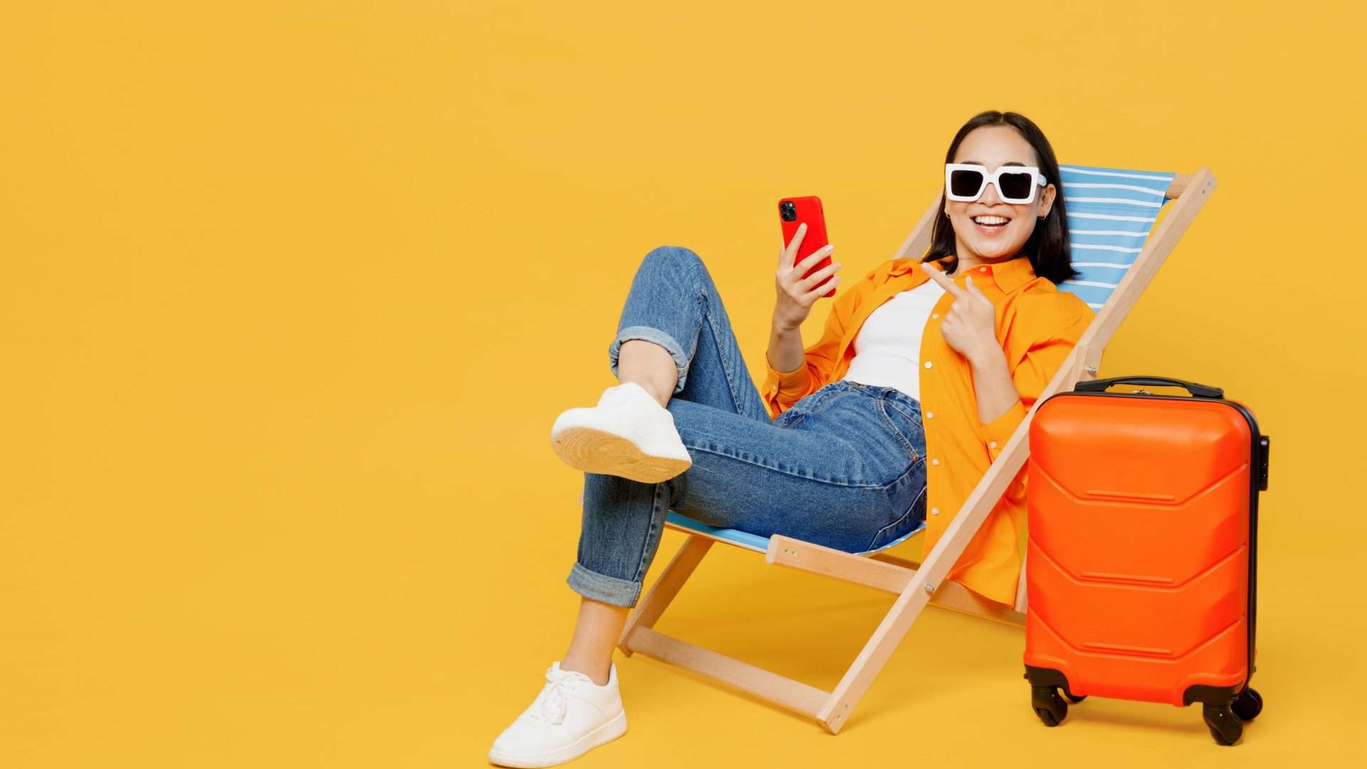 Jovem mulher vestindo roupas de verão sentada em uma espreguiçadeira usando o celular isolada em um fundo amarelo liso. Turista viajando para o exterior no tempo livre para descansar e relaxar. Conceito de viagem de avião.