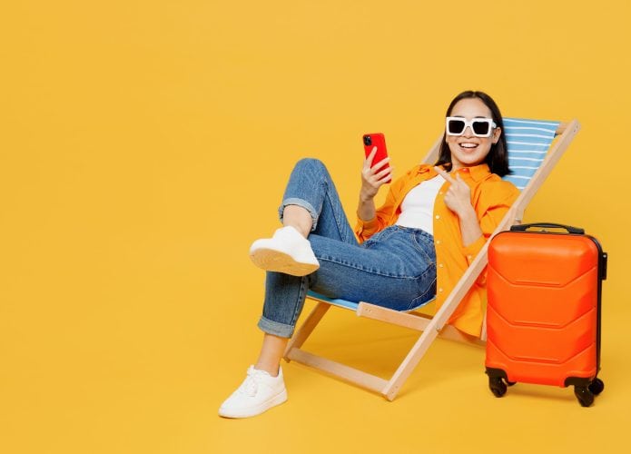 Jovem mulher vestindo roupas de verão sentada em uma espreguiçadeira usando o celular isolada em um fundo amarelo liso. Turista viajando para o exterior no tempo livre para descansar e relaxar. Conceito de viagem de avião.