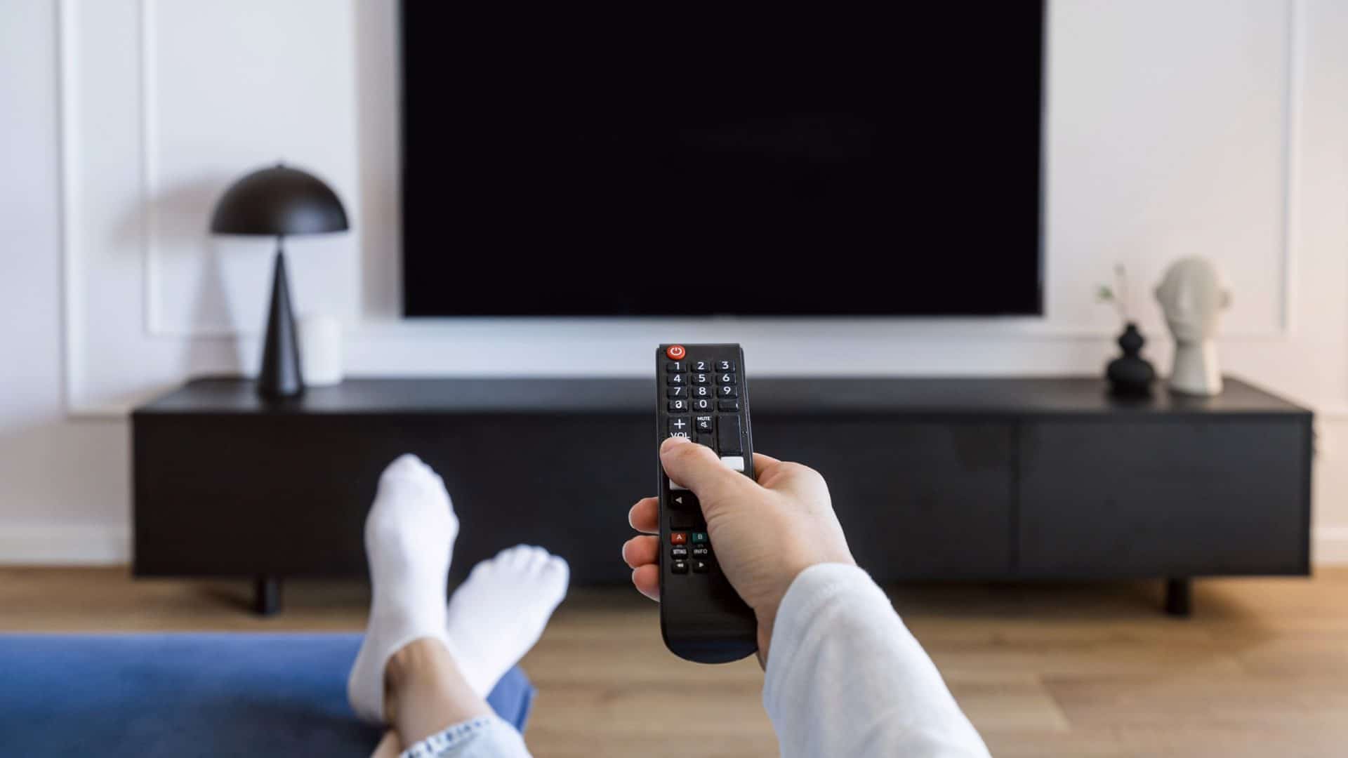 Foco em mão feminina com controle remoto, com pés para cima, e TV desligada ao fundo