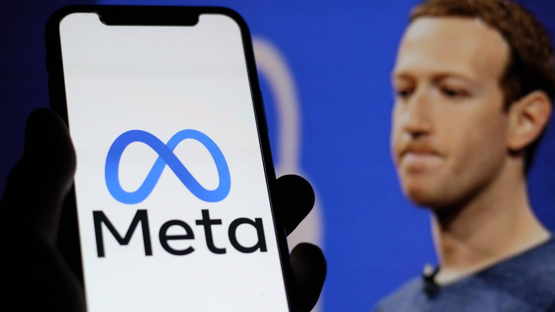 Celular com logomarca da Meta na tela e, ao fundo, Mark Zuckerberg com rosto franzido