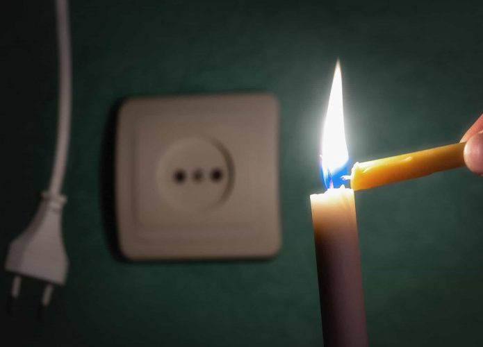 Ao fundo, um plugue de tomada desconectado e uma tomada ao lado; à frente, em foco, uma vela sendo acesa