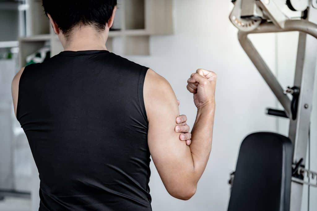 Imagem mostra um homem de costas na academia. Ele está vestindo uma regata preta e segura o bíceps, como se estivesse com dor.