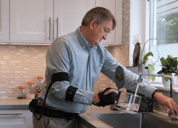 Imagem mostra um homem idoso segurando uma chaleira embaixo do jato de água da pia. Ele usa uma luva robótica na mão direta.