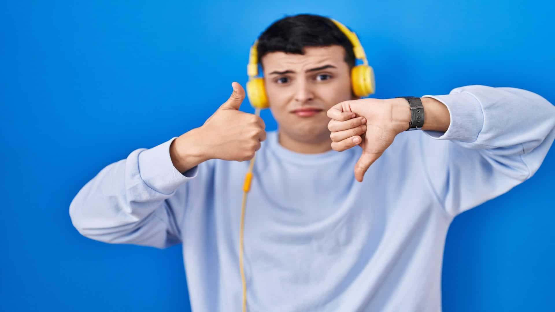 Pessoa não binária ouvindo música usando fones de ouvido fazendo polegares para cima e para baixo, expressão de desacordo e concordância. conflito louco