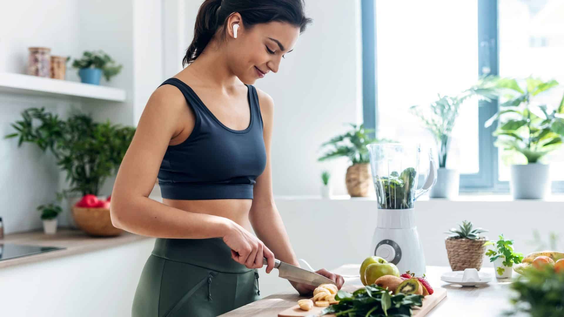 Corta de uma mulher atlética preparando frutas e legumes para fazer um smoothie enquanto escuta música com fones de ouvido na cozinha de casa.