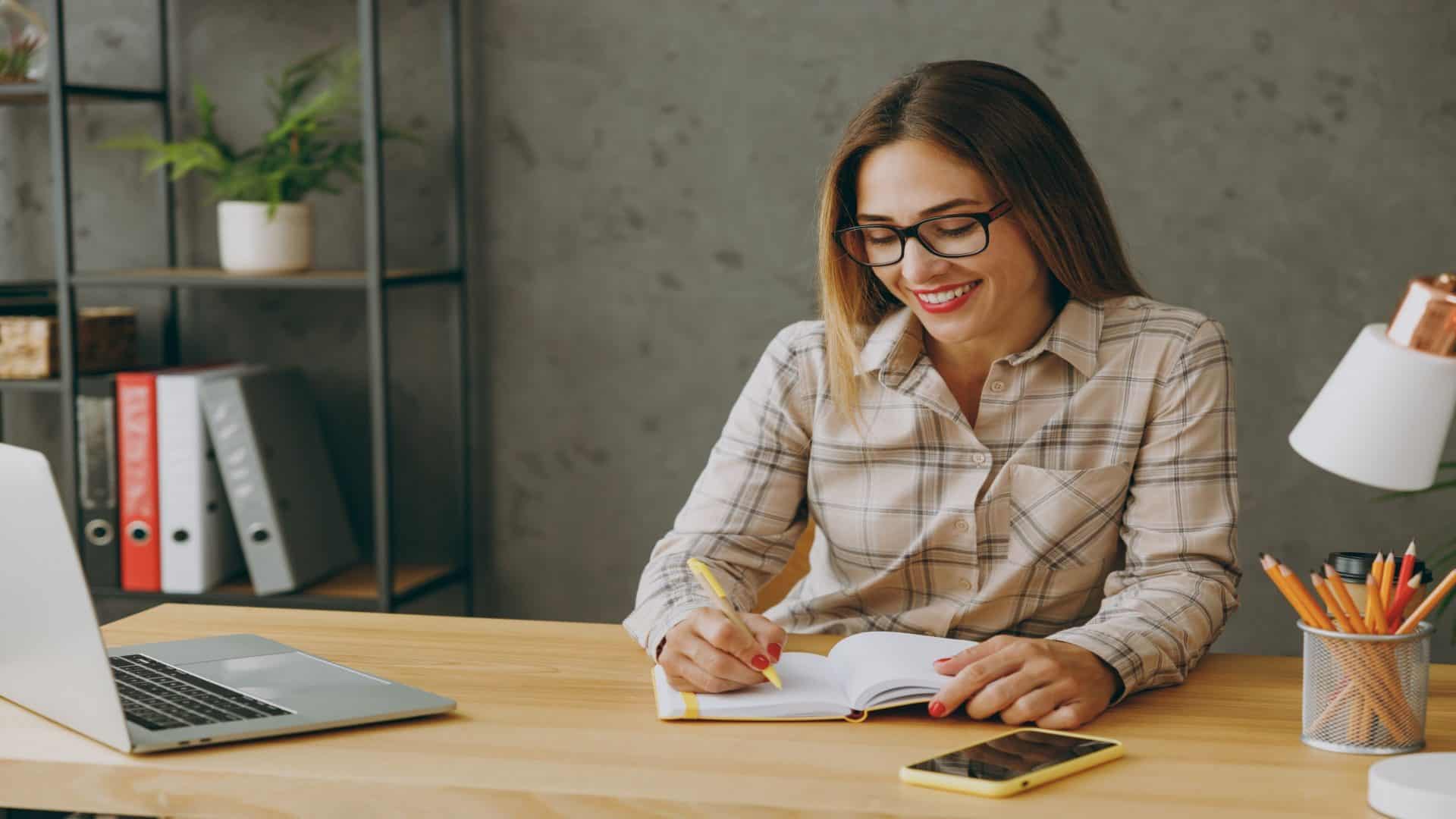 Empregada jovem e animada, vestindo uma camisa e roupas casuais, com óculos, sentada na mesa do escritório com um laptop, olhando para a câmera e anotando algo em um caderno, fazendo algumas anotações. Conceito de conquista na carreira.