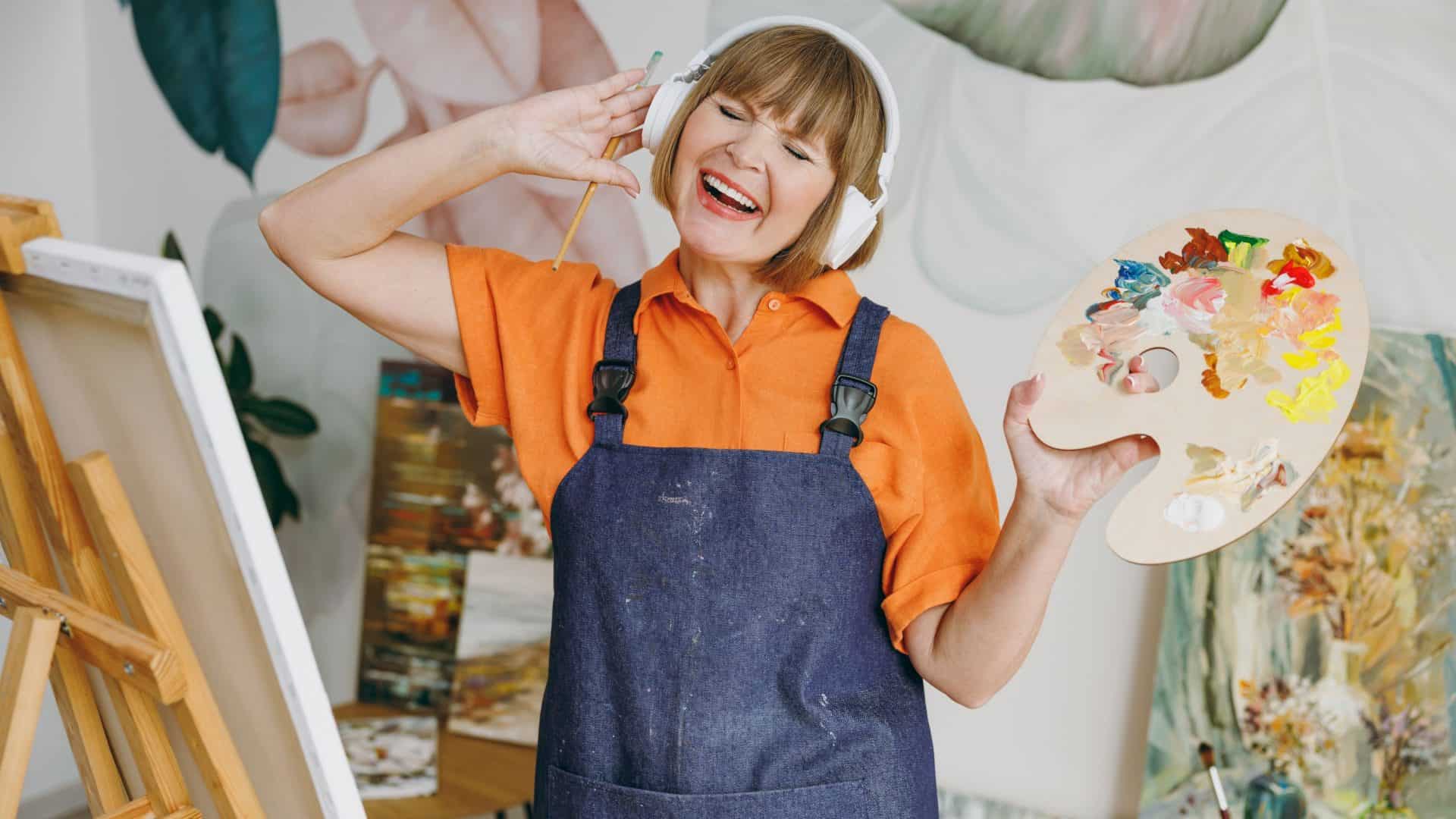 Artista divertida idosa de 50 anos, vestindo roupas casuais, está perto de um cavalete com uma pintura, ouvindo música de fones de ouvido e passando o tempo livre na sala de estar. Conceito de lazer e hobby.