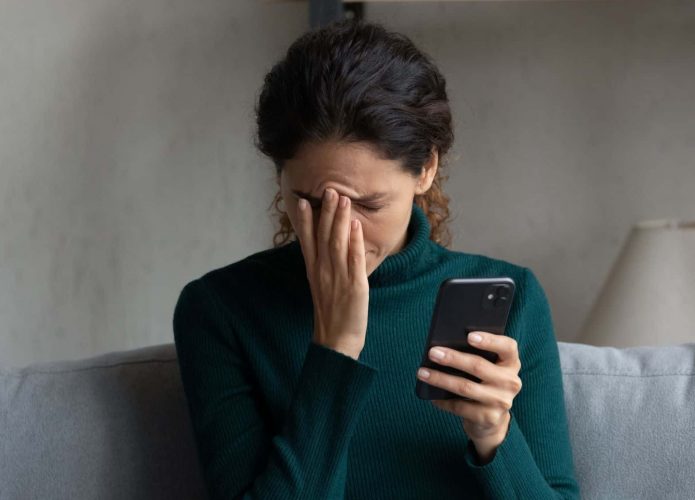 Mulher com mão no rosto e expressão preocupada enquanto segura celular