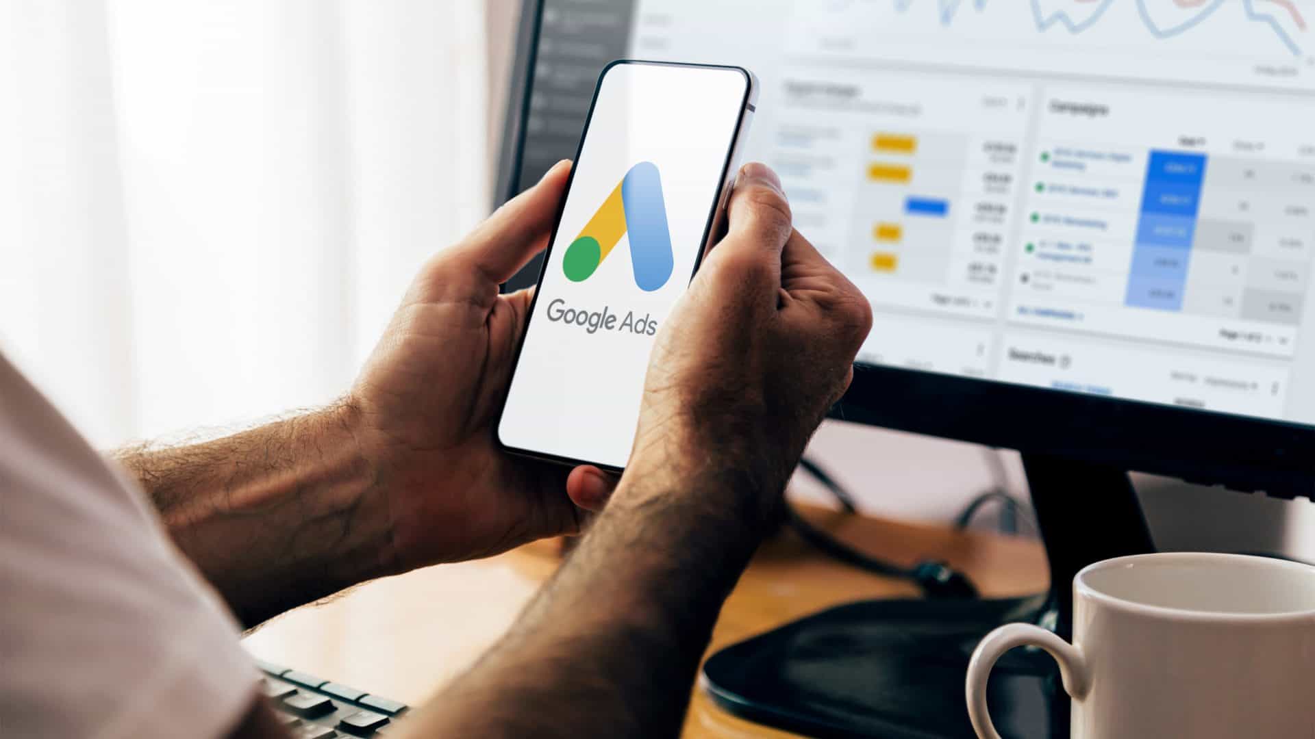 Homem segurando celular com logotipo do Google Ads na tela e, ao fundo , computador com gráficos no monitor