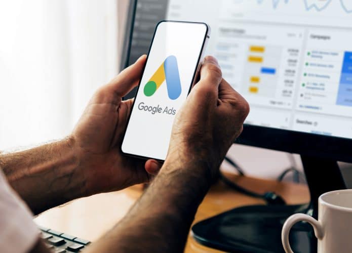 Homem segurando celular com logotipo do Google Ads na tela e, ao fundo , computador com gráficos no monitor