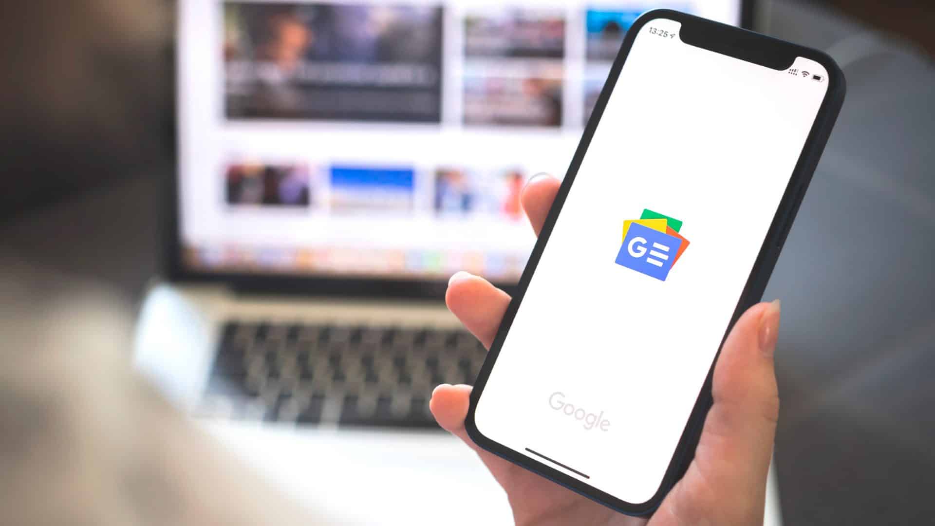 Pessoa segurando iPhone com logomarca do Google notícias enquanto agregador de notícias está aberto num Macbook atrás