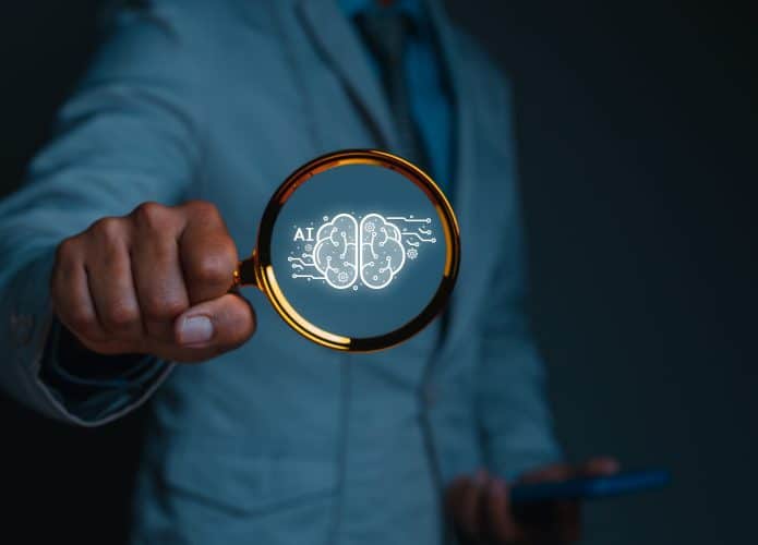 Pessoa segurando lupa com ilustração de cérebro e sigla de inteligência artificial