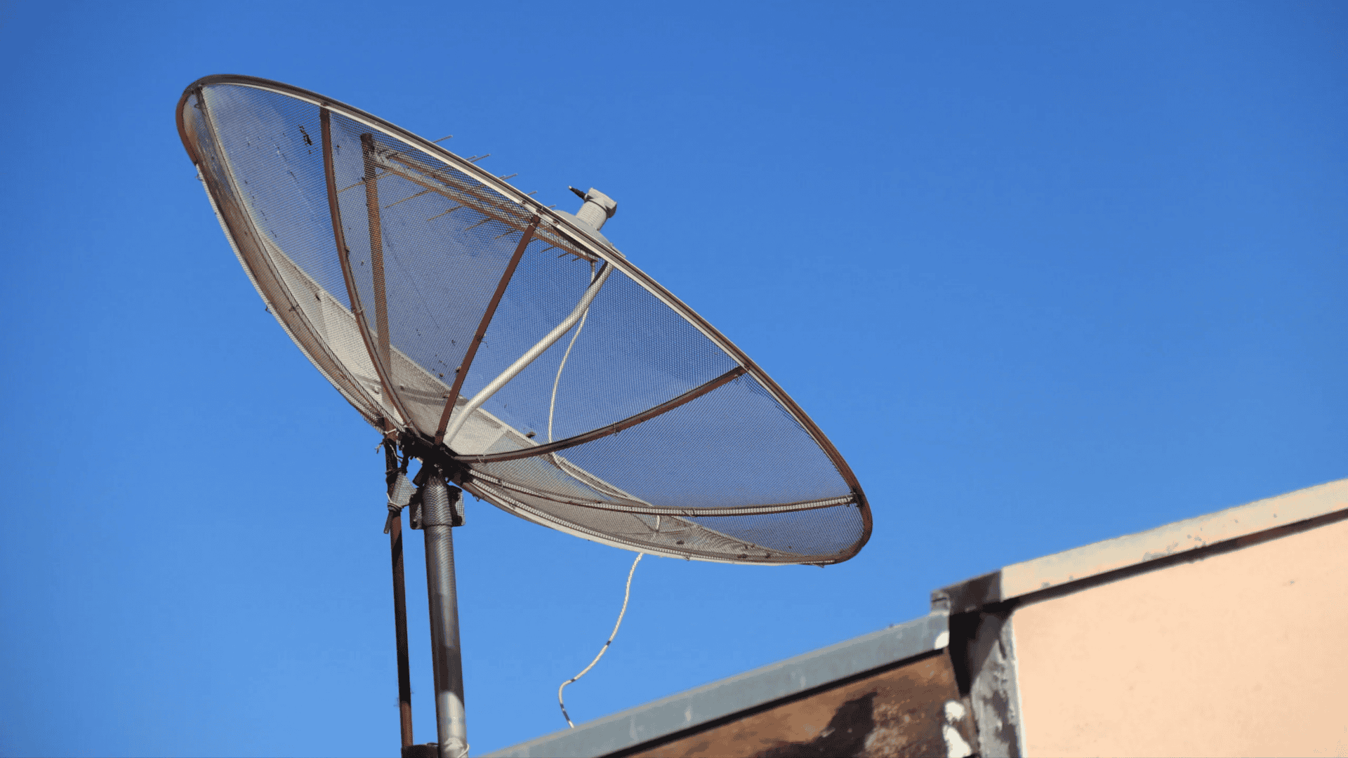 imagem mostra uma antena parabólica antiga em cima de um telhado com um céu azul límpido no fundo