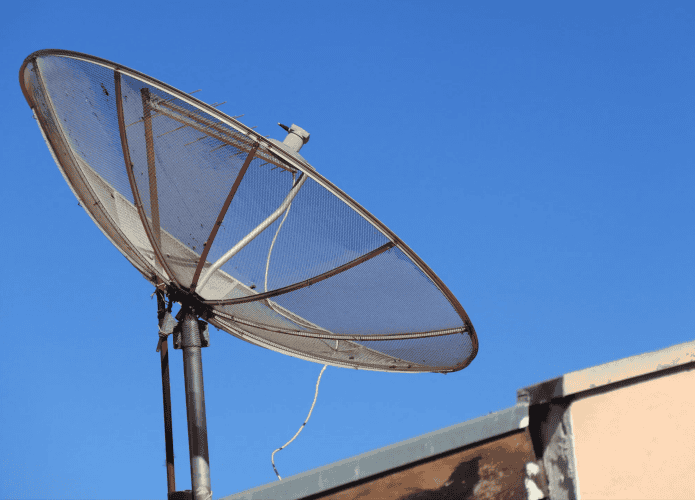 imagem mostra uma antena parabólica antiga em cima de um telhado com um céu azul límpido no fundo