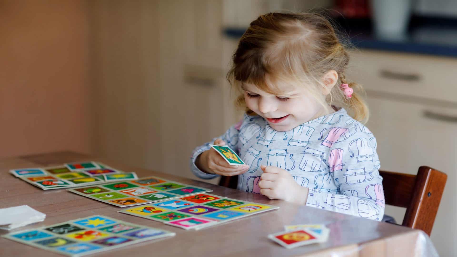 Garotinha fofa e sorridente brincando de um jogo de cartas. Criança feliz e saudável treinando a memória, pensando. Lazer e educação criativa em casa. Atividade em família.