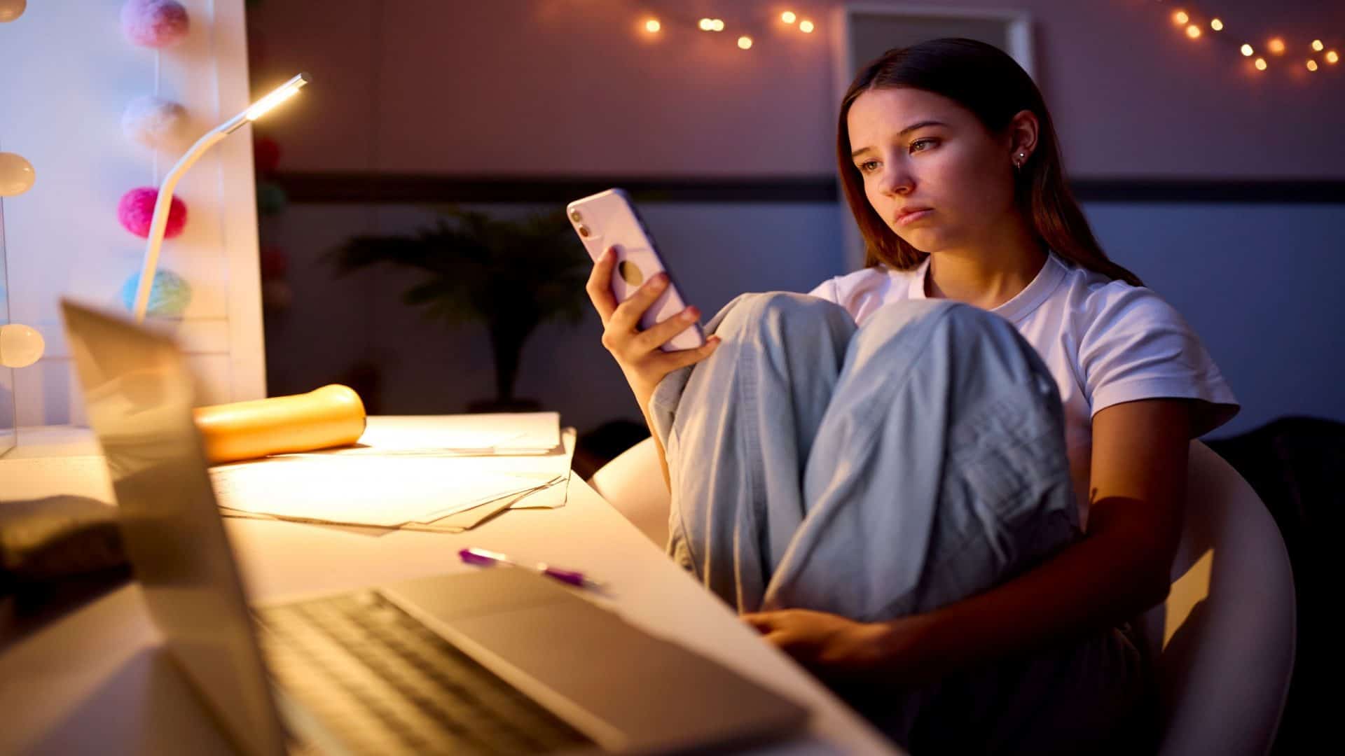 Menina adolescente preocupada sentada à mesa no quarto em casa olhando para o celular à noite.