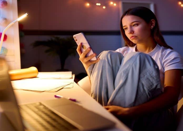 Menina adolescente preocupada sentada à mesa no quarto em casa olhando para o celular à noite.