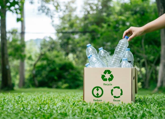 Imagem mostra um jardim em uma área externa. Em primeiro plano, à direita, há uma caixão de papelão com o símbolo da reciclagem e garrafas plásticas vazias no interior