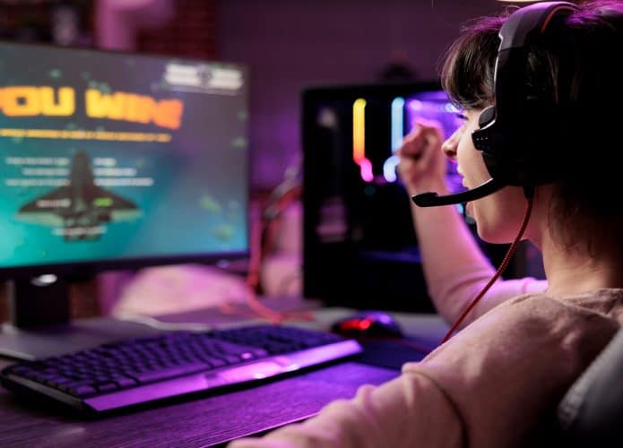 Jogadora empolgada comemorando a vitória em um duelo de videogame multiplayer online de naves espaciais, derrotando os oponentes. Mulher aproveitando seu tempo livre em um apartamento com luzes RGB, se sentindo nas nuvens com a vitória.