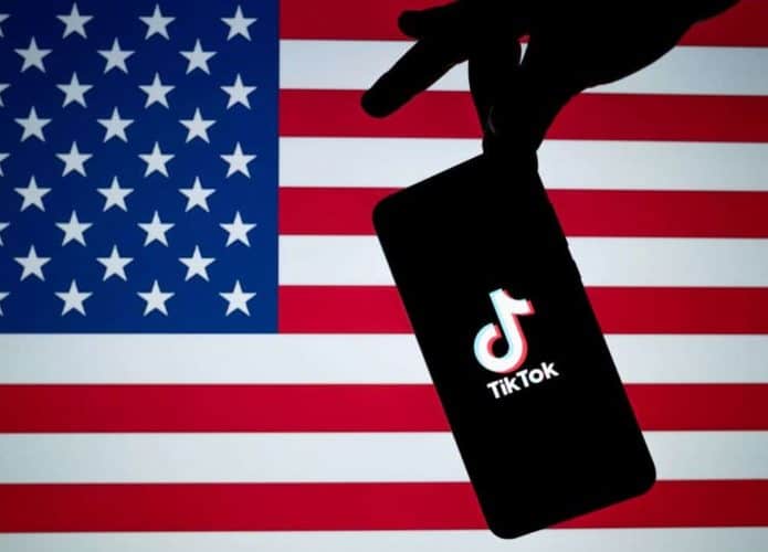 Pessoa segurando, com a ponta dos dedos de maneira parecida a uma pinça, um celular com a logomarca do TikTok na tela na frente de uma bandeira dos Estados Unidos
