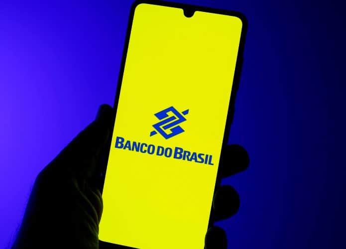Pessoa segurando celular em frente fundo azulado com logomarca do Banco do Brasil na tela