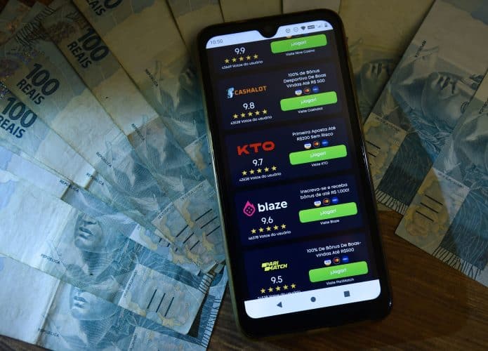 Celular com lista de bets que funcionam no Brasil em cima de notas de 100 reais