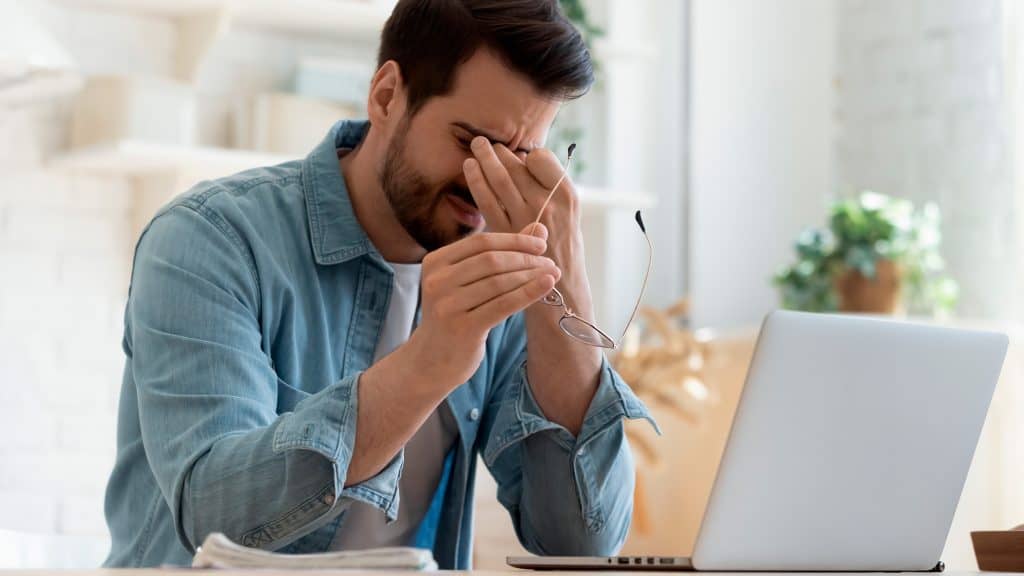 Homem com enxaqueca segurando óculos enquanto usa notebook