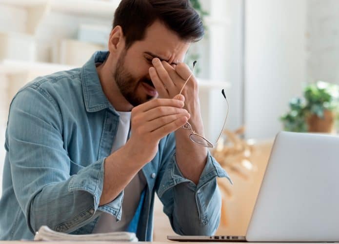 Homem com enxaqueca segurando óculos enquanto usa notebook