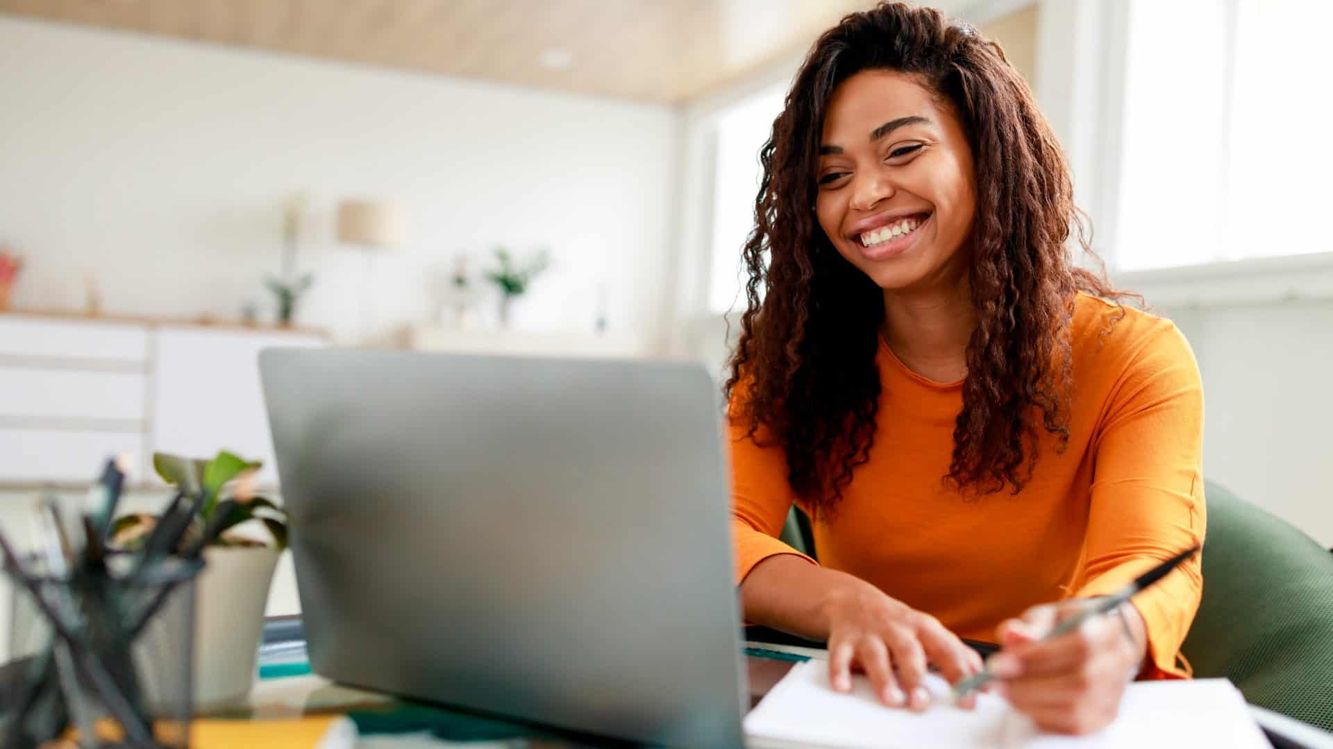 Mulher fazendo anotações em caderno enquanto olha sorrindo para notebook