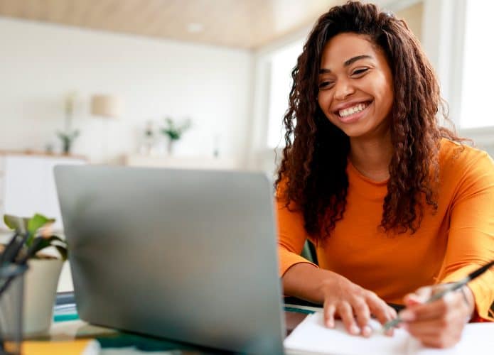 Mulher fazendo anotações em caderno enquanto olha sorrindo para notebook