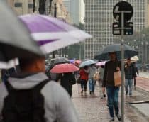 Pessoas agasalhadas e carregando guarda-chuvas andando em avenida de São Paulo