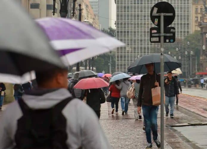 Pessoas agasalhadas e carregando guarda-chuvas andando em avenida de São Paulo