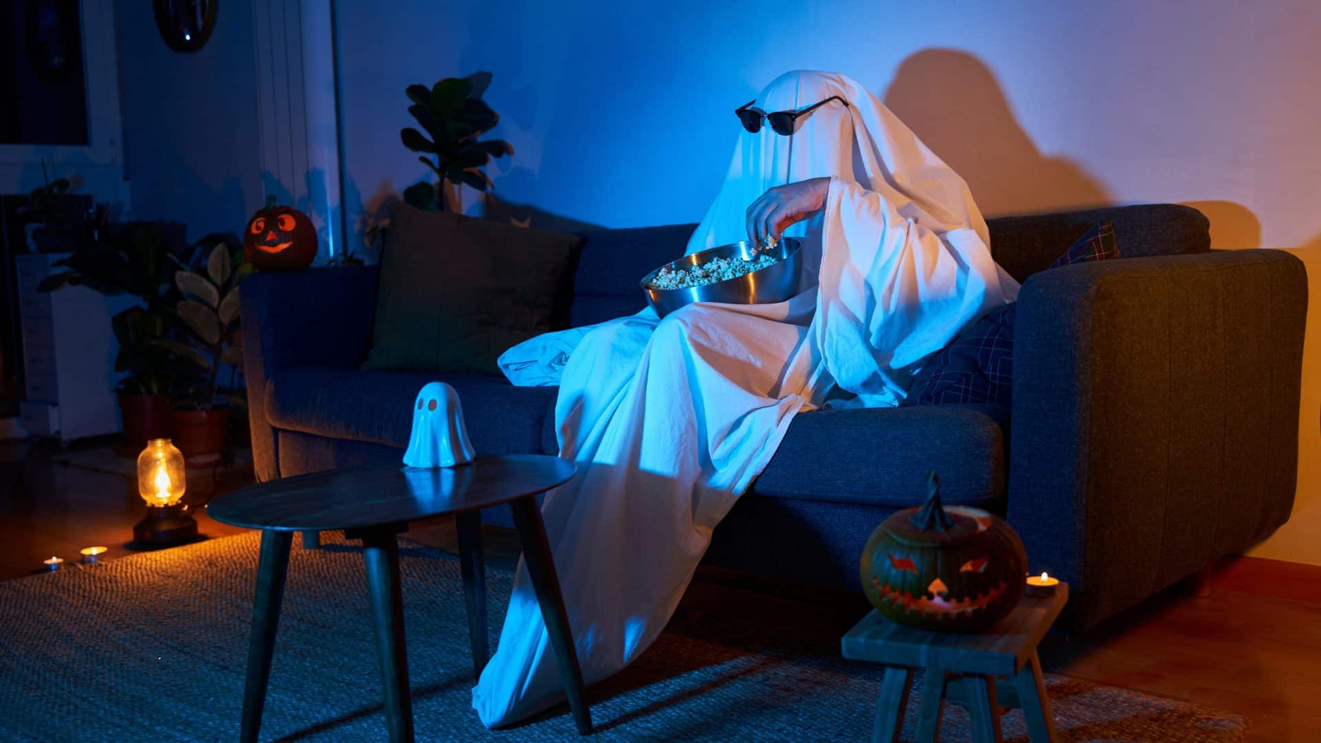 Pessoa fantasiada de fantasma com óculos escuros sentada no sofá comendo pipoca e assistindo filme