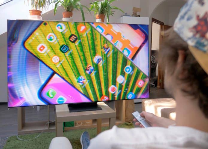 Perspectiva sobre ombro de moço sentado assistindo vídeo sobre celulares em TV da Samsung