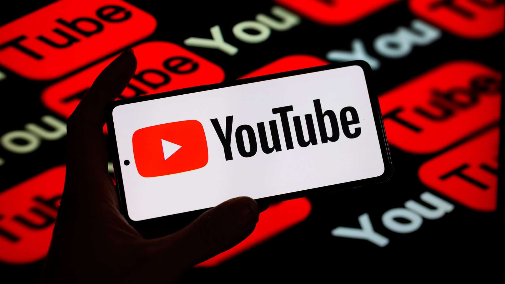 Pessoa segurando celular com logomarca do YouTube na tela na frente de outra tela com logomarcas do YouTube também
