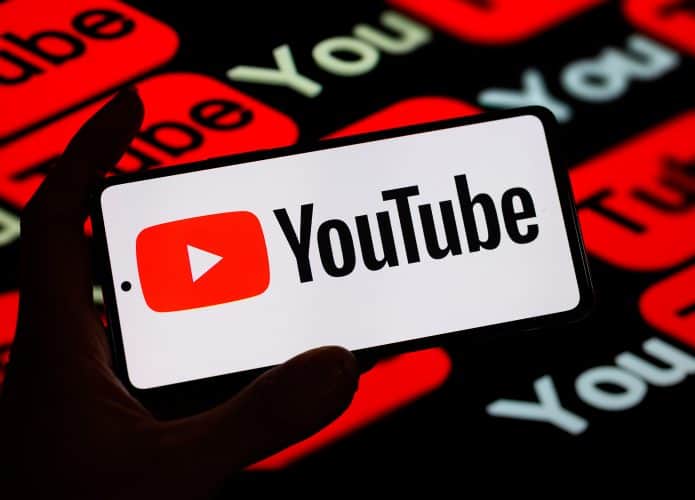 Pessoa segurando celular com logomarca do YouTube na tela na frente de outra tela com logomarcas do YouTube também