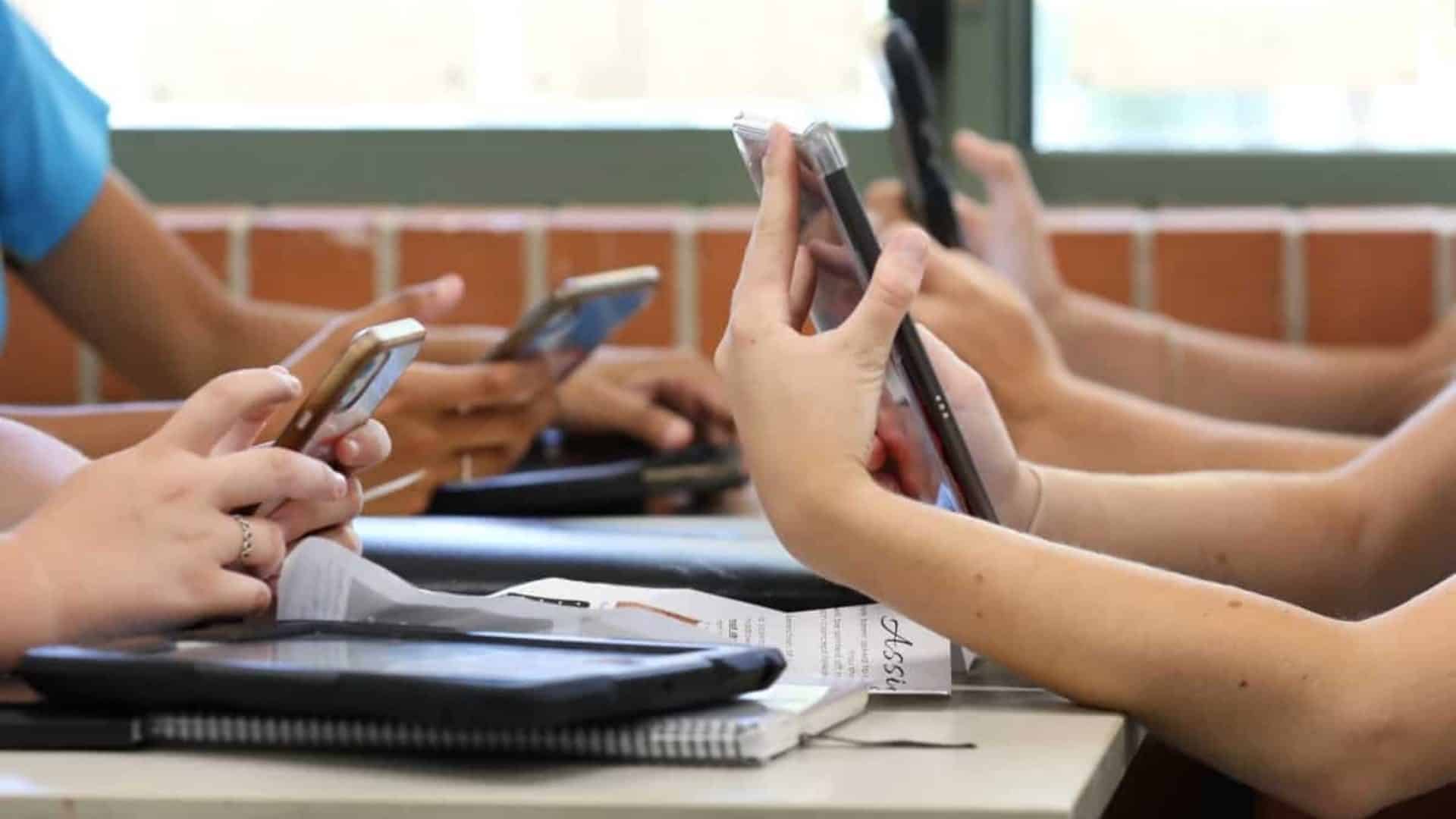 Mesa com alunos do ensino médio, todos segurando vários dispositivos digitais, como telefones e tablets, durante uma aula