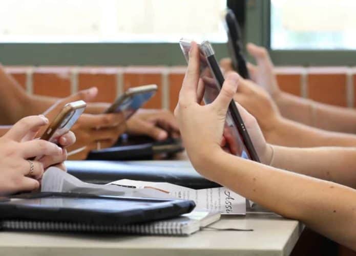 Mesa com alunos do ensino médio, todos segurando vários dispositivos digitais, como telefones e tablets, durante uma aula
