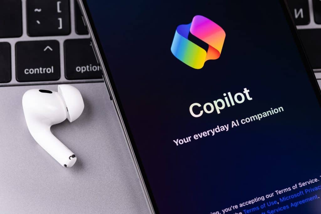 Logo do Copilot em um smartphone, que se encontra em cima de um notebook e ao lado de um fone de ouvido