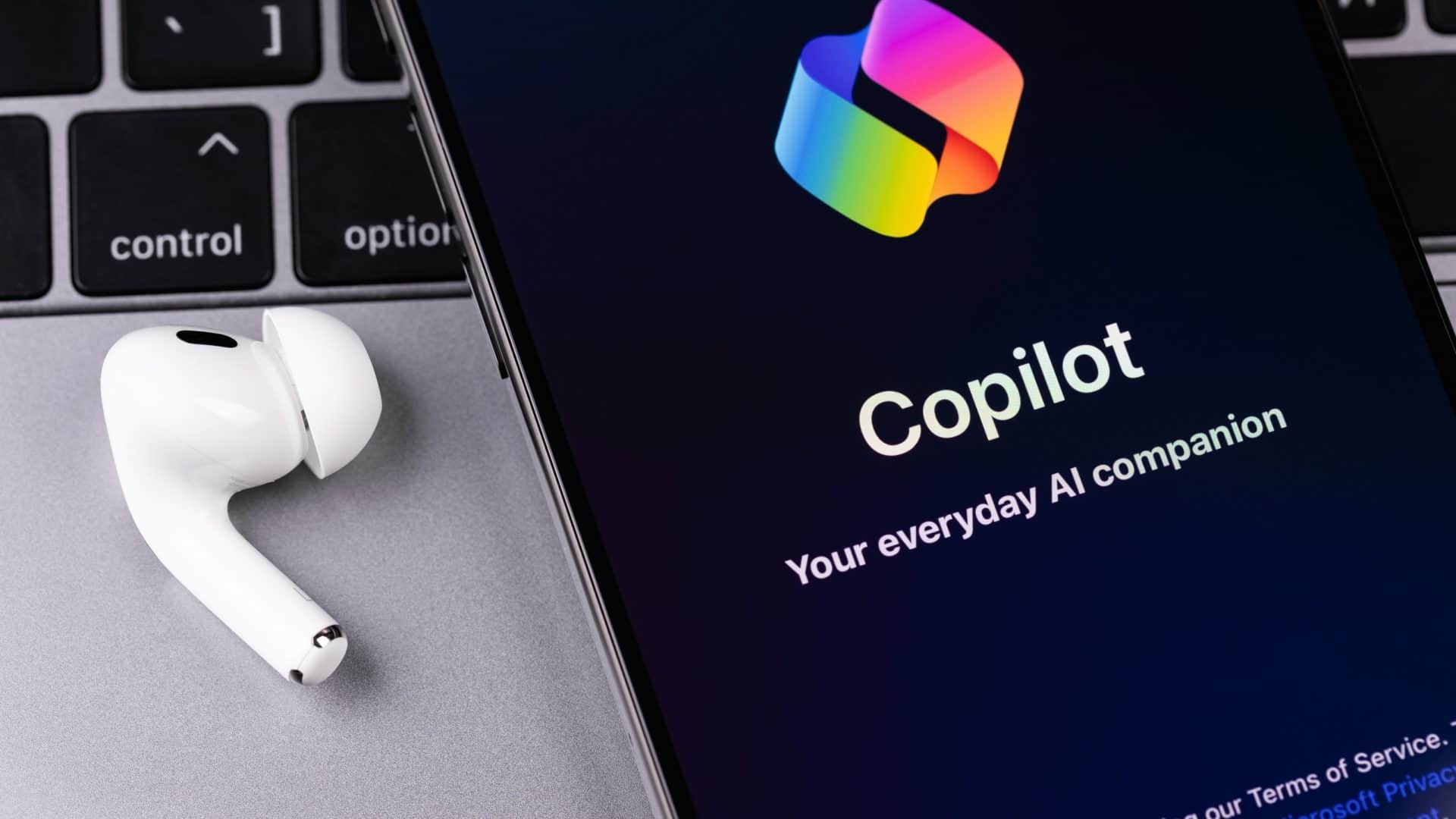 Logo do Copilot em um smartphone, que se encontra em cima de um notebook e ao lado de um fone de ouvido