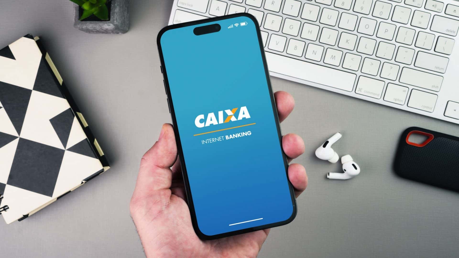 Homem segurando um smartphone com o aplicativo do banco Caixa Econômica Federal na tela sobre uma mesa de fundo cinza.