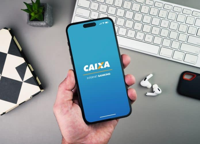 Homem segurando um smartphone com o aplicativo do banco Caixa Econômica Federal na tela sobre uma mesa de fundo cinza.