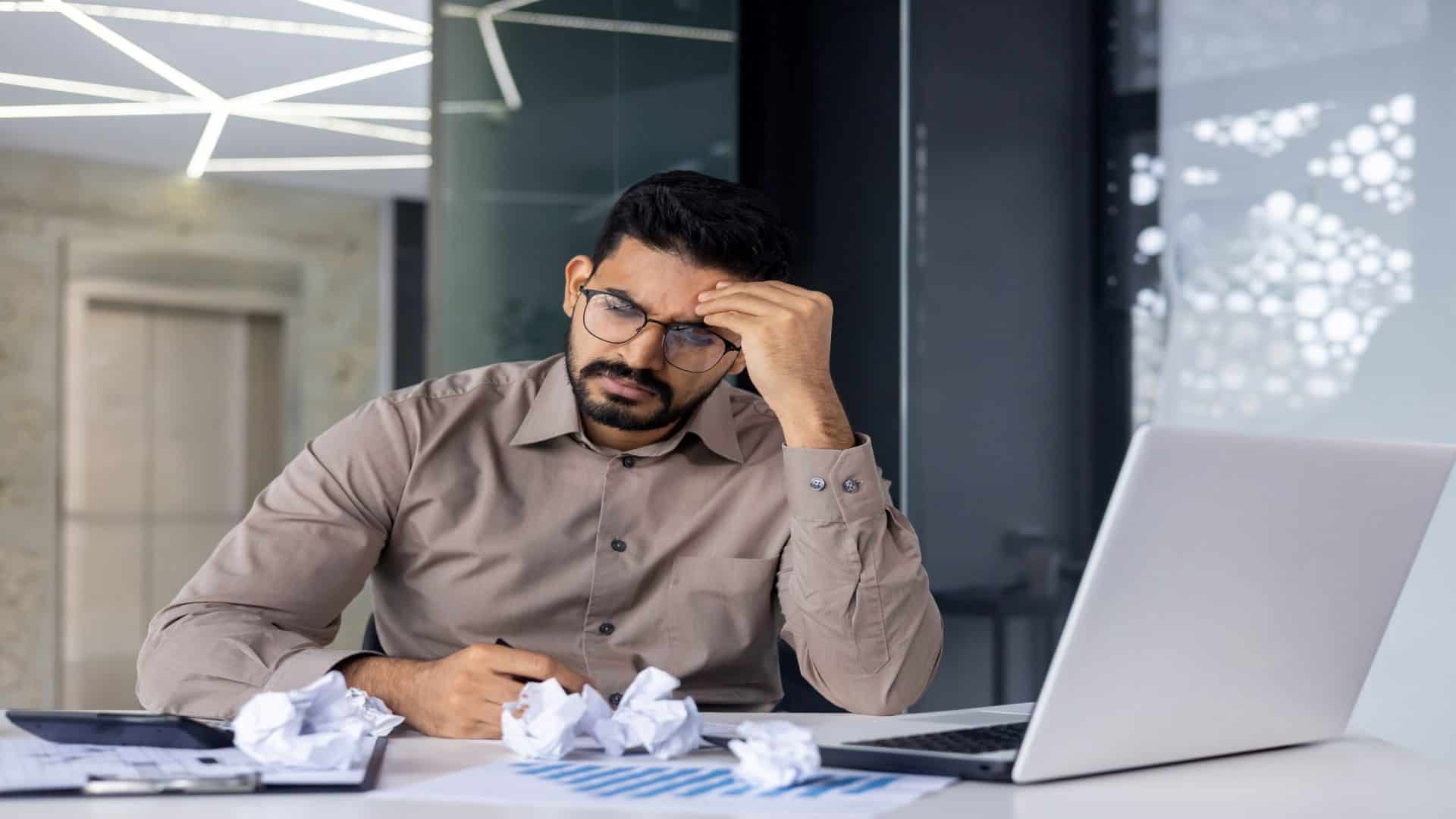 Empresário desapontado, triste e depressivo, sentado entre papéis e documentos amassados dentro do escritório, homem insatisfeito com os resultados, relatórios financeiros ruins, contador infeliz