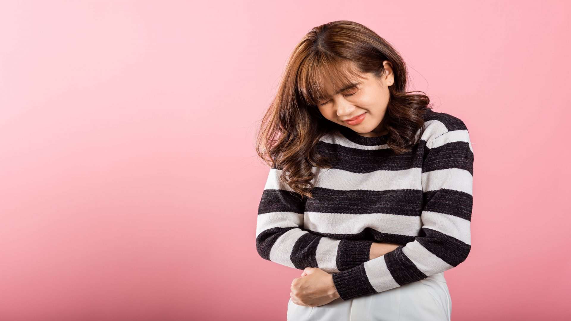 Retrato de uma mulher asiática sentindo dor de estômago e abdominal em uma foto de estúdio isolada em um fundo rosa. Representando o conceito de saúde e medicina da gastrite.