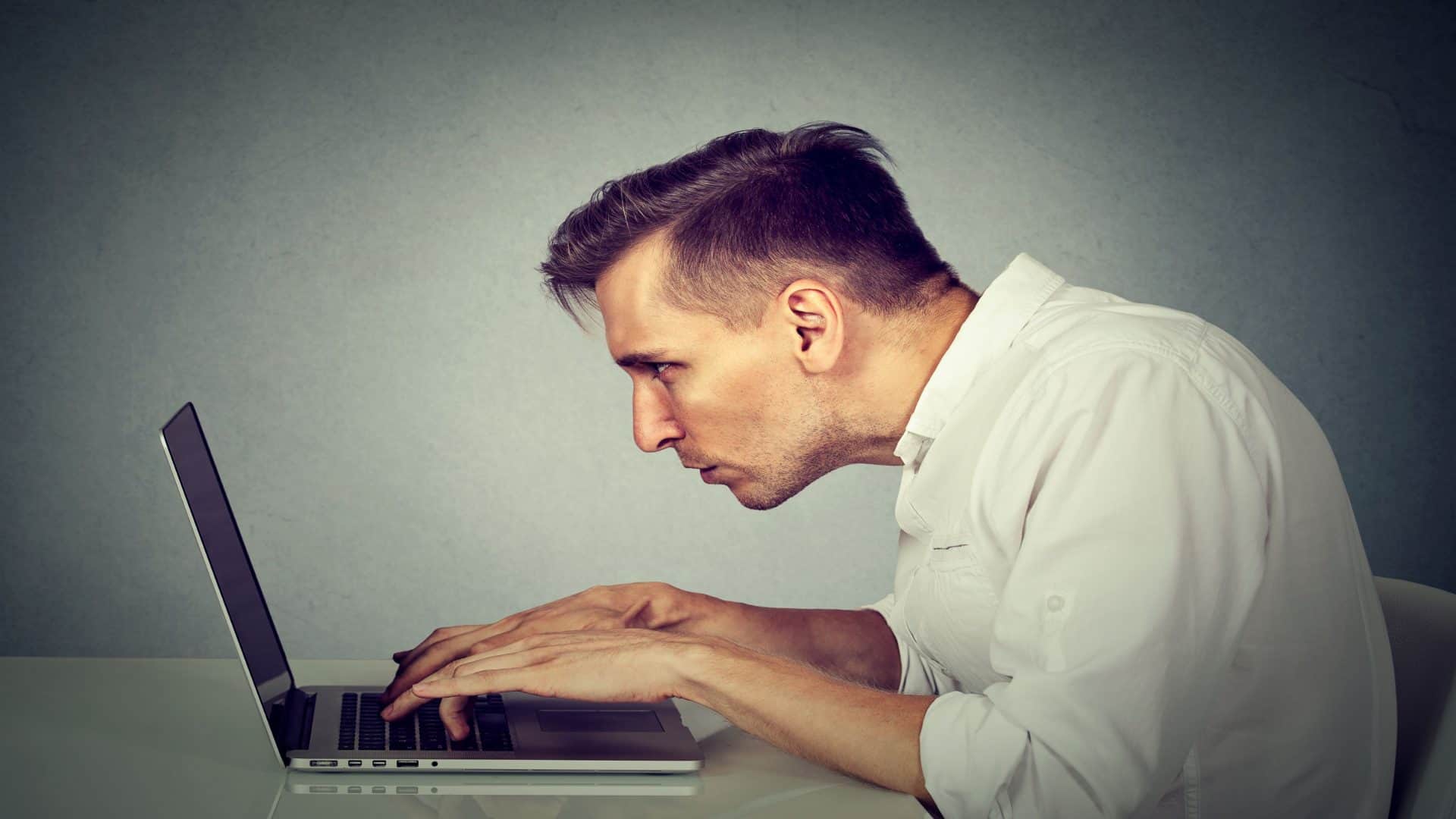 Perfil lateral de um jovem trabalhando no computador, sentado à mesa, isolado no fundo do escritório com parede cinza. Conceito de uma vida longa, monótona e cansativa de horas de trabalho.