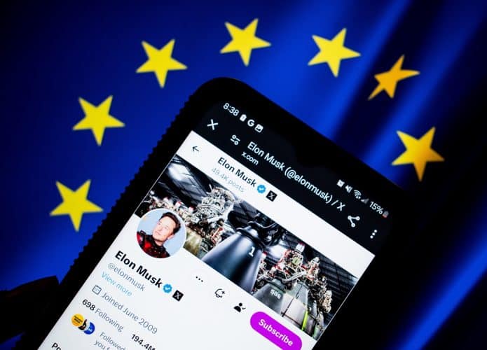 Bandeira da União Europeia no fundo; à frente, smartphone exibindo o perfil de Elon Musk no X